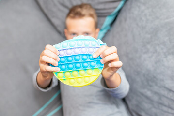 Naklejka premium Schoolboy 10s boy lying on the couch and playing with sensory pop it fidget toy at home. Kid playes stress and anxiety relief fidgeting game after learning.