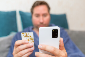 Young man buying fashion items through the Internet on cellphone by using credit card.Online Payment. Frontal portrait of man holding debit card and using smartphone for shopping at home