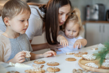 Mom and children decorate Christmas gingerbread at home. A boy and a girl paint with cornets with sugar icing on cookies. New Year's decor, branches of a Christmas tree.