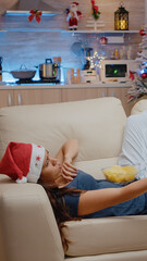 Bored adult switching channels with TV remote control while laying on couch. Woman wearing santa hat and watching television, eating chips from bowl on christmas eve festivity.