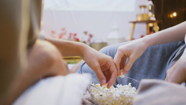 Lovely couple joins hands and takes delicious popcorn from bowl on pillows in room with large fabric as screen on wall at romantic date close backside view