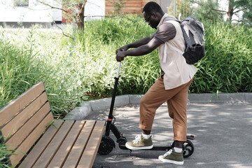 Horizontal side view long shot of young African American guy wearing casual outfit with backpack parking electric scooter near wooden bench