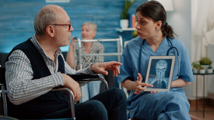 Fototapeta na wymiar Medical assistant holding tablet with x ray scan for checkup with disabled man in nursing home. Nurse explaining radiography on digital device to senior patient sitting in wheelchair