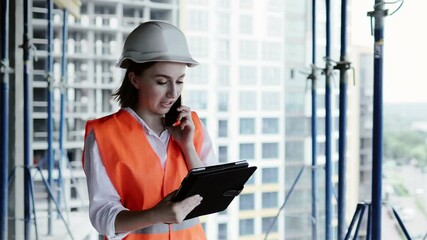 Young female civil engineer in safety jacket and helmet is talking on mobile phone on development construction site outdoor while working on laptop.