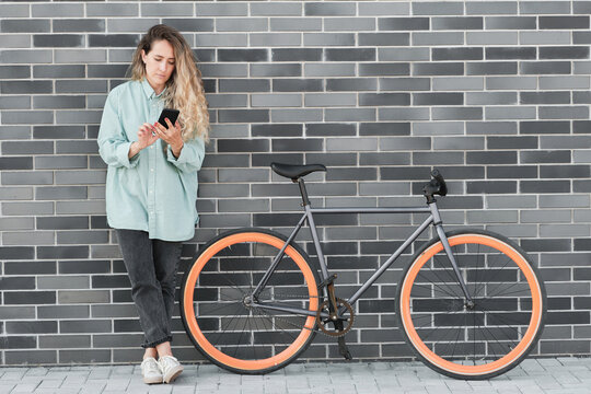 Young Caucasian woman leaning against gray brick wall outdoors with bicycle parked next to her watching something in Internet on smartphone