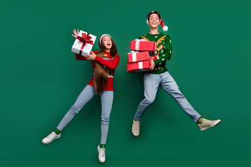 Photo of inspired two friends jump hold stack giftboxes wear santa hat ugly pullover isolated green color background