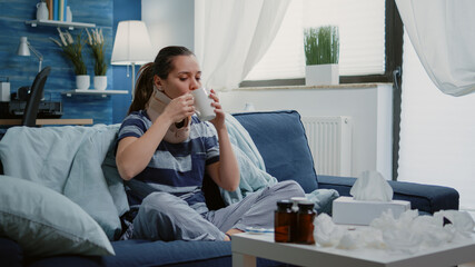 Adult with cold and flu wearing cervical neck collar while sitting on sofa. Ill woman with virus...