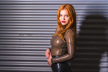 Redhead girl in latex, tight brown and black suit, on a metal wall - obrazy, fototapety, plakaty