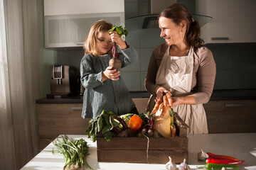 mom and child daughter in a linen apron with a box of fresh garden vegetables in the kitchen. Organic Concept, clean eating 