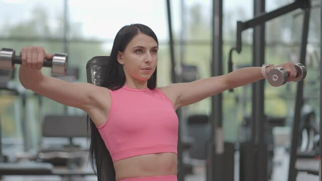 A brunette woman in a pink suit raises dumbbells to the sides while training her shoulders in the gym. Seated Shoulder and Arm Bench Exercise