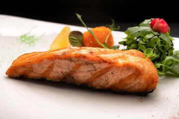Fried salmon steak with herbs and lemon on a white plate on a dark background