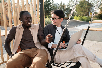 Horizontal medium portrait of modern African American man and Asian woman sitting together on swing...