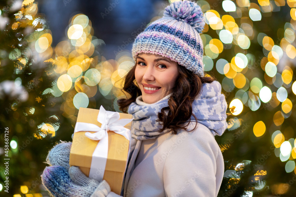 Poster winter holidays, celebration and people concept - happy smiling woman with christmas gift over festive lights