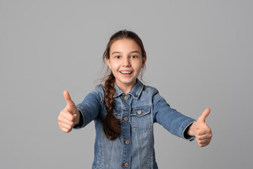 happy smiling young girl in casual clothes looking at camera and showing thumbs up, isolated on grey background. Positive emotions