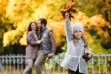 Happy family in autumn park. Young parents of little preschool girl hugging during walk outdoors in forest with colorful leaves on trees. Joyful daughter and caring mom and dad together on weekend