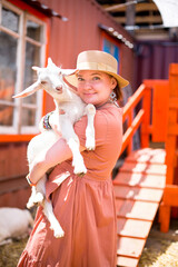 fat racy girl in a hat and dress on the farm holding a baby goat in her arms