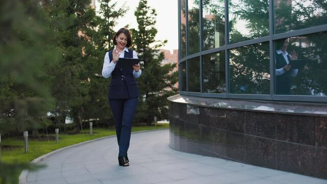 A businesswoman walks slowly down the street and communicates via Skype. The girl uses a tablet for video communication with colleagues. Beautiful cityscape of the business center.