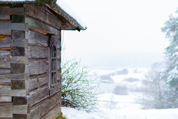 the wall, roof and window of an old gray wooden log house against the background of a snow-covered landscape and a forest in the distance