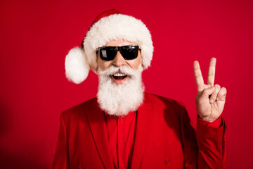 Photo of friendly carefree santa claus show v-sign toothy smile wear sunglass x-mas hat suit on red color background