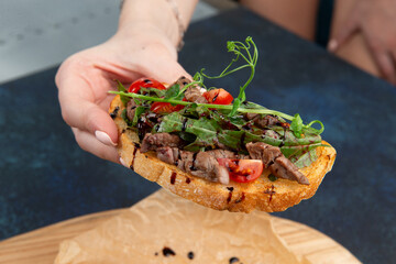 Hand holds a bruschetta with meat, tomatoes and herbs. Against a dark background.