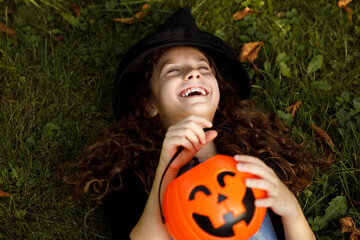 Cute little girl laughing in a witch costume, lying on the grass, holding a cauldron in her hand.