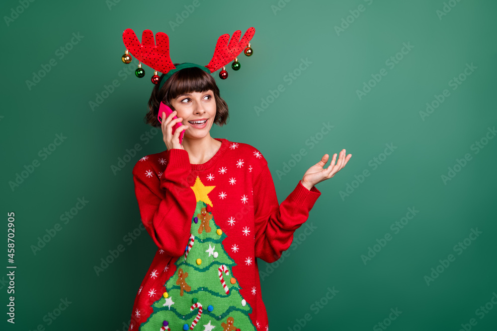 Wall mural Photo of cute charming woman dressed red pullover antlers talking modern device looing empty space isolated green color background