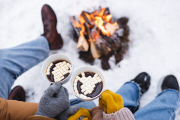 Cropped view of couple in gloves holding cups with marshmallows near blurred bonfire in winter park