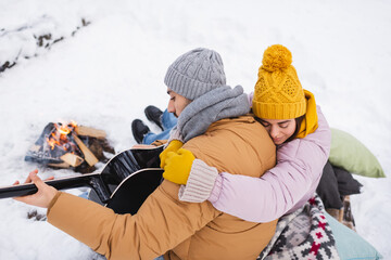 High angle view of woman hugging boyfriend playing acoustic guitar near bonfire in winter park