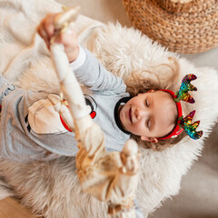 Beautiful happy girl child with Christmas deer antlers in fashionable pajamas plays with a toy and lies on the bed, top view. Winter Holidays Concept