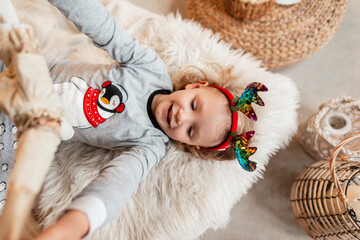 Cheerful beautiful little girl child with a smile and christmas antlers in fashionable pajamas lies on the bed and plays with a toy, top view