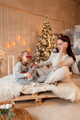 Beautiful young woman mom with a cute baby girl in fashion clothes sit on the bed and have fun near Christmas trees and lights. Winter road family holidays