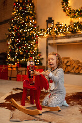 Obraz na płótnie Canvas Happy beautiful little girl child in fashionable pajamas is played with a nutcracker and a red swing horse near the fireplace on the background of Christmas decorations