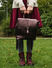 Front view of female legs in grey tights and skirt. Stylish burgundy shoes. Leather bag in the arms. Green grass. Autumn fashion.