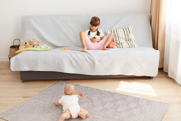 Two sisters: little schoolgirl sitting on sofa and toddler baby lying on carpet play with popit ob floor, children wearing casual closing, spending time at home.