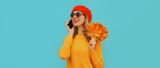 Autumn portrait of happy smiling young woman with yellow maple leaves calling on smartphone wearing a red french beret on blue background