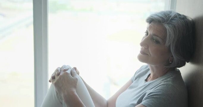 A beautiful senior citizen woman spending all day at the window during her quarantine and self-isolation. Pleasant thoughts about the future help her forget about loneliness and the elderly.
