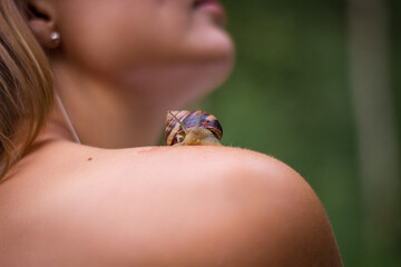 Obraz premium A snail sits on the shoulder of a Caucasian woman. On a close-up of the skin, the mollusk is clearly visible. The girl's face with a seashell against the background of a green forest.