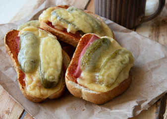 Hot sandwiches made of white bread, ham, pickles and cheese on a sheet of craft paper on a wooden background with a brown mug close-up