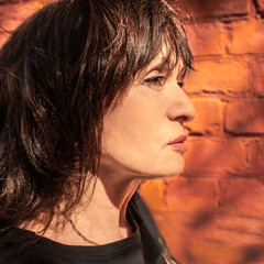 Profile of a 65 year old brunette woman against a warm tone brick wall background.
