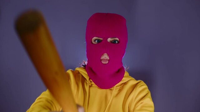Young woman in pink balaclava with baseball bat in hands. Hooligan in mask beats with bat, looking at camera.