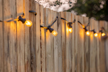 Cosy light bulbs lined up in a row, against a wooden garden fence. There are some green bushes and green grass in the background. It's a sunny Summer day evening at a country wedding or other