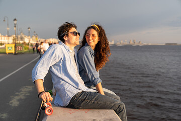 Stylish young couple sitting on concrete pier at seaside enjoy sunset. Happy man and woman in...