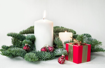Christmas composition with wrapped red gift box, lighted candles and glittering red baubles surrounded with fir branches on white background.