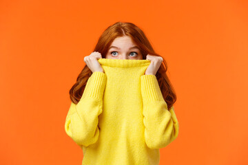 Scared cute teenage redhead girl pulling sweater on face and peeking at upper left corner with...