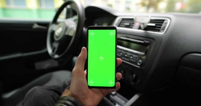 Male hand holding empty green screen chroma key of smart phone in the car mockup