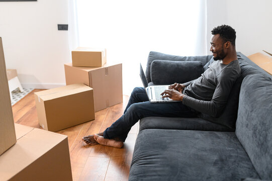 Side view at satisfied African-American guy sitting on the comfortable couch and using laptop computer among cardboard boxes, smiling black man moving into new apartment, shopping online, web surfing