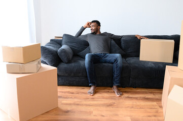 Satisfied African-American guy sitting on the comfortable couch and looks away dreamly among cardboard boxes, smiling black man moving into new apartment, relocated in new house, rent a flat