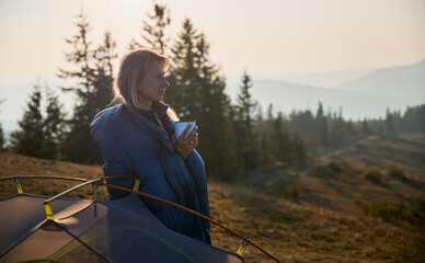 Side view of female tourist in blue sleeping bag looking thoughtfully at the scenery in front of...