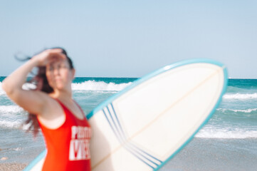 Surfer girl on the beach holding surf board watching ocean waves in sunny day. Adult woman prepare to surfing. Summer vacation lifestyle.