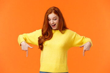 Wondered and excited, amused cute redhead woman in yellow sweater pointing fingers down, look...